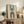 May include: Framed geometric art print with cream background featuring two arch shapes - blue striped arch at top, brown striped arch below with teal center circle, and "BAUHAUS 1923" text vertically arranged on right side. Displayed on rustic wooden sideboard alongside fern plant, books, and ceramic vase in neutral-toned living room with beige armchair.