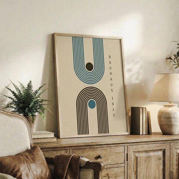 May include: Framed geometric art print with cream background featuring two arch shapes - blue striped arch at top, brown striped arch below with teal center circle, and "BAUHAUS 1923" text vertically arranged on right side. Displayed on rustic wooden sideboard alongside fern plant, books, and ceramic vase in neutral-toned living room with beige armchair.
