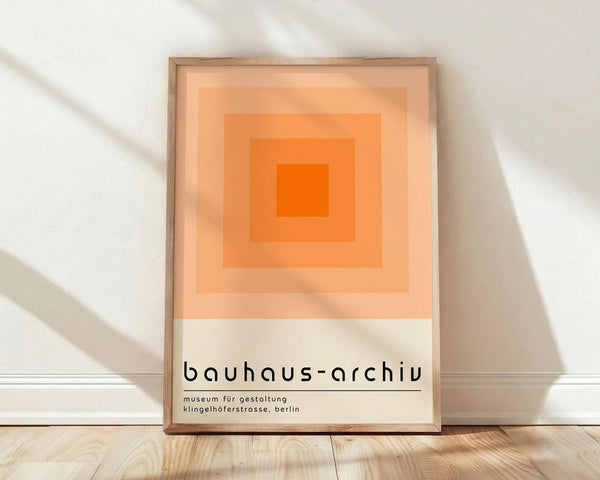 May include: Framed poster with orange gradient squares in concentric pattern, transitioning from bright orange center to pale peach edges, with white bottom section containing black text reading "bauhaus-archiv" and museum details. Natural wood frame leaning against white textured wall on light wooden floor with soft window light shadows.