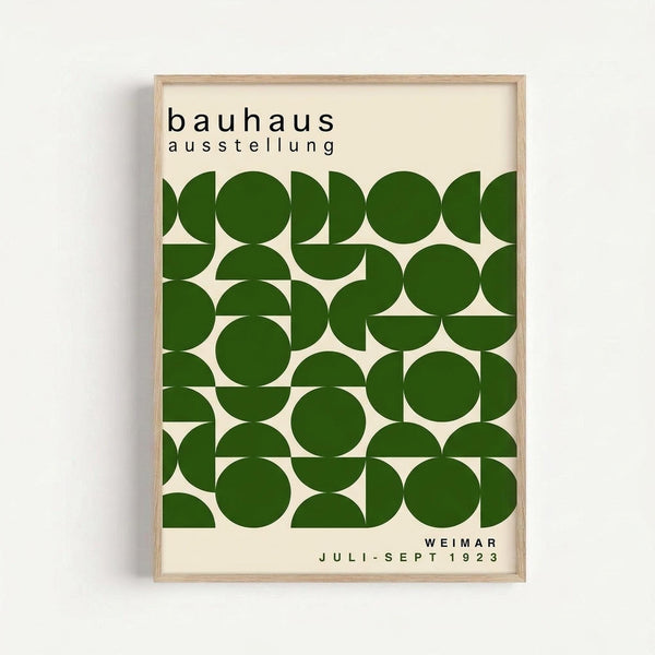 Framed poster with cream background featuring geometric green circles and semicircles arranged in a grid pattern. Black text at top reads "bauhaus ausstellung" and bottom shows "WEIMAR JULI-SEPT 1923". The artwork uses bold forest green shapes creating a modernist, Bauhaus-style design. Light wood frame mounted on white wall.