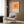 May include: Modern living room with beige armchair and geometric pillow, brass floor lamp, dark side table with open book, and wood wall panels. Orange gradient squares poster with Bauhaus Archiv Berlin text hangs in white frame above chair. Neutral tones with warm lighting and minimalist mid-century modern decor styling.