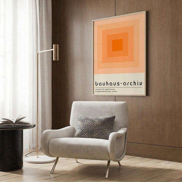May include: Modern living room with beige armchair and geometric pillow, brass floor lamp, dark side table with open book, and wood wall panels. Orange gradient squares poster with Bauhaus Archiv Berlin text hangs in white frame above chair. Neutral tones with warm lighting and minimalist mid-century modern decor styling.
