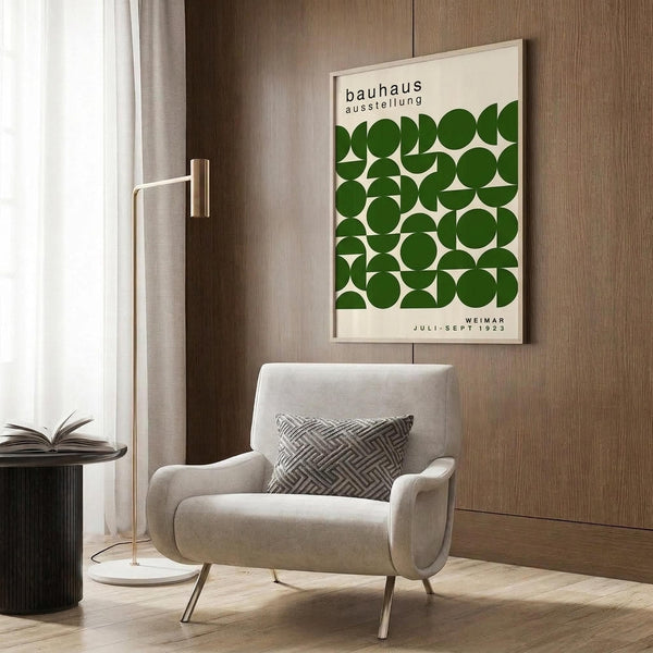 May include: Framed Bauhaus exhibition poster with green geometric circles and semicircles on cream background, displayed above beige upholstered armchair with patterned pillow. Modern living room setting features brass floor lamp, dark round side table with open book, wood panel wall, and white curtains. Poster shows "Weimar Juli-Sept 1923" text.