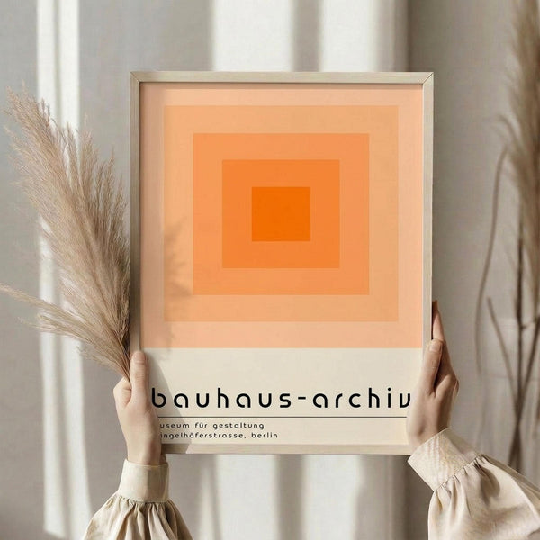 May include: Person holding framed poster featuring concentric orange squares in gradient shades from deep orange center to pale peach outer rings, with black text below reading "bauhaus-archiv" and museum details. Natural wood frame, neutral beige setting with dried pampas grass visible to left side.