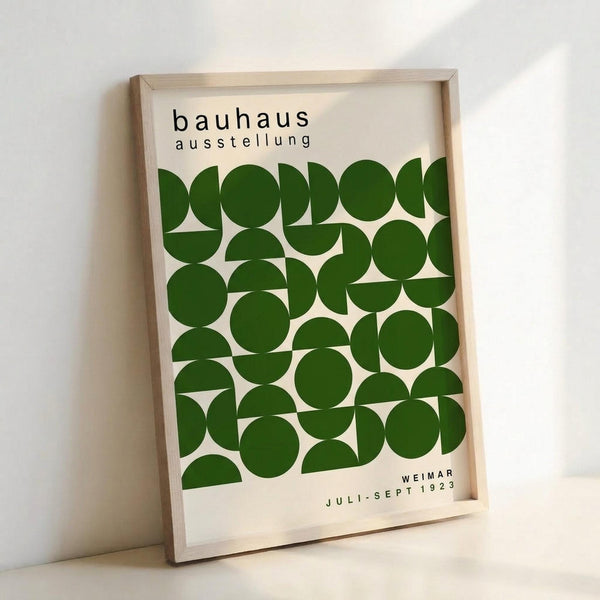 May include: Framed poster with cream background featuring geometric pattern of green circles and semicircles arranged in rows. Black text reads "bauhaus ausstellung" at top and "WEIMAR JULI-SEPT 1923" at bottom. Light wood frame leans against white wall with soft shadow, displaying minimalist Bauhaus exhibition design aesthetic.