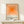 May include: Framed poster with orange gradient squares design and "bauhaus-archiv" text displayed on light wood console table against white wall. Small wrapped gift with evergreen sprig sits nearby. Minimalist Scandinavian-style interior setting with natural lighting creating soft shadows.