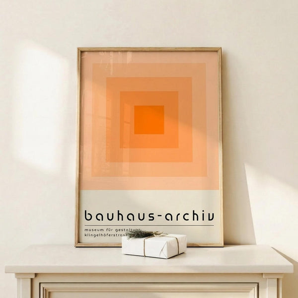 May include: Framed poster with orange gradient squares design and "bauhaus-archiv" text displayed on light wood console table against white wall. Small wrapped gift with evergreen sprig sits nearby. Minimalist Scandinavian-style interior setting with natural lighting creating soft shadows.