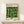 May include: Framed Bauhaus exhibition poster with geometric green circles and semicircles arranged in a grid pattern on cream background, displayed on white wooden console table next to small wrapped gift with evergreen sprig, against neutral wall with natural lighting