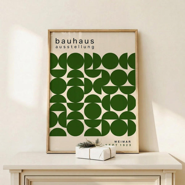 May include: Framed Bauhaus exhibition poster with geometric green circles and semicircles arranged in a grid pattern on cream background, displayed on white wooden console table next to small wrapped gift with evergreen sprig, against neutral wall with natural lighting