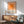 May include: Orange geometric poster with concentric squares in graduating shades from deep orange center to pale cream edges, hung in wooden frame above mid-century modern credenza. Sage green upholstered armchair with wooden arms sits beside round coffee table. White ceramic table lamp and natural sunlight streaming across white walls complete the minimalist room setting.