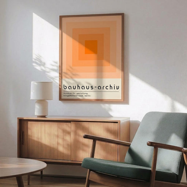 May include: Orange geometric poster with concentric squares in graduating shades from deep orange center to pale cream edges, hung in wooden frame above mid-century modern credenza. Sage green upholstered armchair with wooden arms sits beside round coffee table. White ceramic table lamp and natural sunlight streaming across white walls complete the minimalist room setting.
