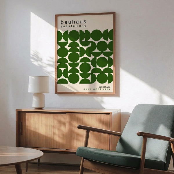 May include: Framed vintage Bauhaus exhibition poster with green geometric circles on cream background, displayed above mid-century modern wood credenza with white ceramic lamp. Mint green upholstered armchair with wooden arms sits nearby on wooden floor with natural window light creating shadows on white wall.