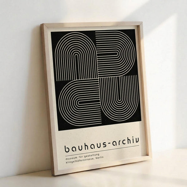 Framed poster with black background featuring white geometric line art forming interconnected rounded rectangular shapes in a grid pattern. Below the artwork, black text reads "bauhaus-archiv" followed by smaller German text mentioning "museum für gestaltung" and "klingelhöferstrasse, berlin". The print is displayed in a light wood frame against a white wall, leaning at an angle.