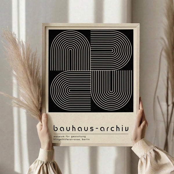 May include: Person in beige ruffled sleeves holding framed black and white geometric poster with concentric linear patterns forming abstract shapes. Natural wood frame against neutral wall with dried pampas grass visible to left side.