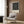May include: Modern beige armchair with geometric patterned pillow in contemporary living room setting. Framed black and white geometric art poster with curved linear design and text mounted on wood-paneled wall. Brass floor lamp and dark round side table with open book nearby. Neutral tones throughout with wood flooring and curtained window.