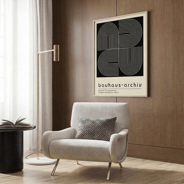 May include: Modern beige armchair with geometric patterned pillow in contemporary living room setting. Framed black and white geometric art poster with curved linear design and text mounted on wood-paneled wall. Brass floor lamp and dark round side table with open book nearby. Neutral tones throughout with wood flooring and curtained window.