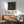 May include: Modern Bauhaus-style poster with black and white geometric line art forming abstract letterforms, displayed in thin wooden frame above mid-century credenza. Scene shows sage green upholstered chair with wooden arms, white ceramic table lamp, and warm natural lighting casting shadows on white wall.