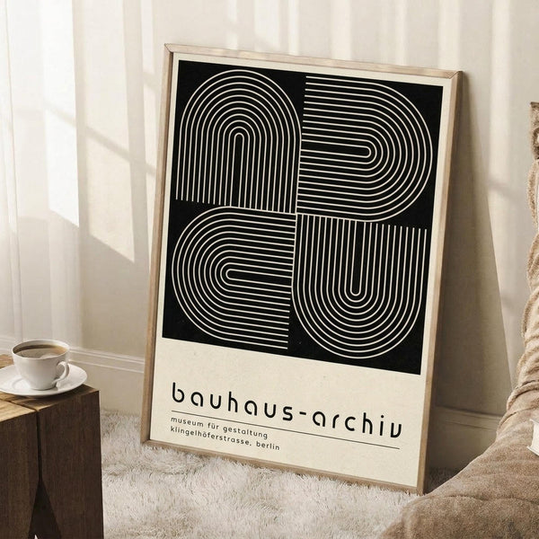 May include: A framed geometric art print leaning against a white wall, featuring black background with white curved parallel lines forming abstract shapes. Light wood frame, displayed beside a coffee cup on wooden block and cream blanket. Natural lighting creates soft shadows on the wall.