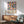 May include: Geometric poster with colorful overlapping circles in pink, blue, orange, and yellow displaying Bauhaus-Archiv museum text, hung above mid-century modern wood sideboard with white ceramic lamp, next to sage green upholstered armchair with wooden frame in sunlit room with white walls