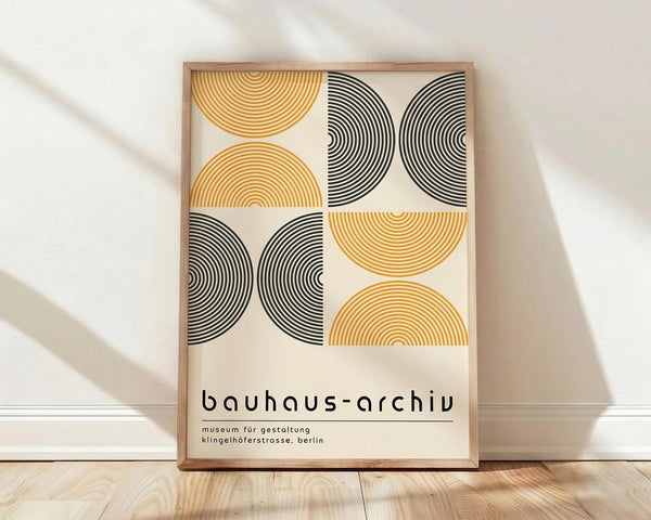 May include: Framed poster with geometric design featuring alternating black and golden yellow semicircular patterns arranged in a grid layout. Clean Bauhaus-style typography below the geometric elements. Light wood frame leaning against white wall on wooden floor with soft natural lighting creating diagonal shadows.