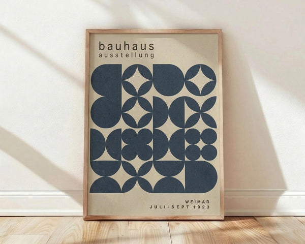 May include: Framed Bauhaus exhibition poster with geometric pattern of overlapping navy blue circles and semicircles on cream background, displayed against white wall on light wood floor with natural window shadows