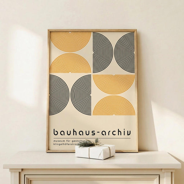 May include: Framed geometric poster with alternating orange and black concentric semicircles in a 2x2 grid pattern, displayed on light wood surface alongside small white gift box with twine and evergreen sprig. Clean minimalist styling with cream background wall.