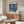 May include: Framed Bauhaus exhibition poster with navy blue geometric circles and shapes on cream background, displayed above wooden credenza in modern living room. White sculptural chair and glass-top coffee table visible. Minimalist Scandinavian-style interior with warm wood tones and neutral palette.