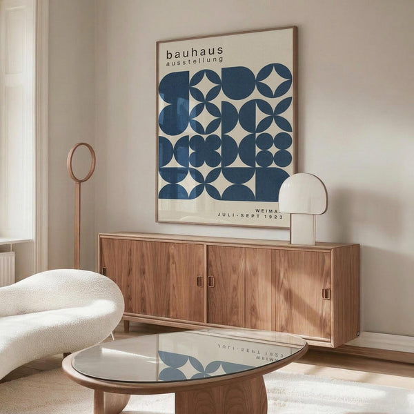 May include: Framed Bauhaus exhibition poster with navy blue geometric circles and shapes on cream background, displayed above wooden credenza in modern living room. White sculptural chair and glass-top coffee table visible. Minimalist Scandinavian-style interior with warm wood tones and neutral palette.