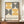 May include: Framed poster with geometric design featuring orange and black concentric semicircles and circles arranged in a grid pattern on cream background, displayed on wooden sideboard next to white ceramic vase and stacked books in elegant room with white wainscoting