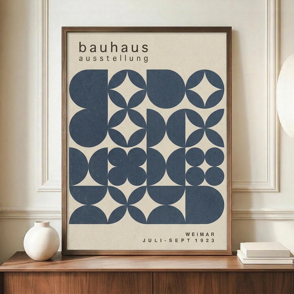 May include: Framed Bauhaus exhibition poster with navy blue geometric circles and overlapping shapes on cream background, displayed on wooden sideboard next to white ceramic vase and stacked books in bright modern room with white paneled walls