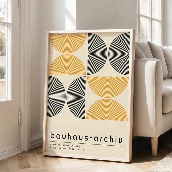 May include: Framed poster with geometric pattern of concentric semicircles in golden yellow and black on cream background, displaying "bauhaus-archiv" text below. Light wood frame leaning against white wall next to beige upholstered sofa in bright, naturally-lit living room with hardwood floors.