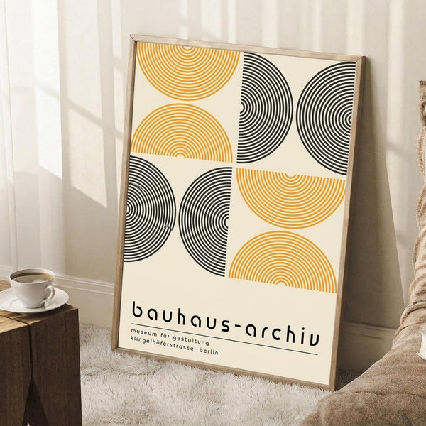 May include: Framed geometric poster with yellow and black concentric semicircles arranged in a grid pattern on cream background, leaning against white wall corner. Small wooden side table with white coffee cup visible beside frame. Beige textured rug and neutral blanket create cozy interior scene with natural lighting.
