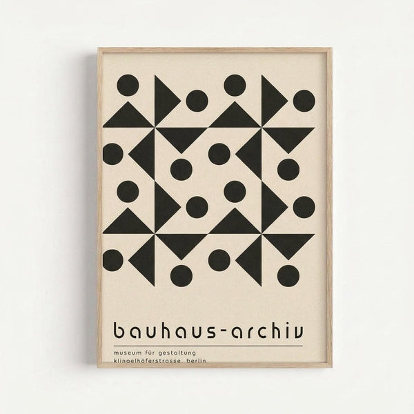 A geometric poster with black triangles and circles arranged in a repeating pattern on cream background, displayed in a light wood frame against a white wall. The minimalist design features four pinwheel-like motifs created from triangular shapes, surrounded by solid circles. Text at bottom reads "bauhaus-archiv" and "museum für gestaltung klinoelhöferstrasse, berlin" in lowercase sans-serif font.