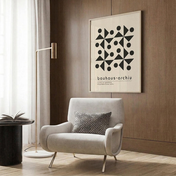 May include: Beige and cream framed Bauhaus poster with black geometric pattern of circles, triangles and diamonds mounted on wood paneled wall above modern light gray armchair with patterned pillow, brass floor lamp and dark round side table with open book in minimalist room setting