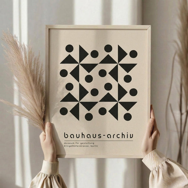 May include: Person in beige sweater holding framed geometric poster with black triangles and circles on cream background, dried pampas grass beside them, neutral curtained window setting, light wood frame