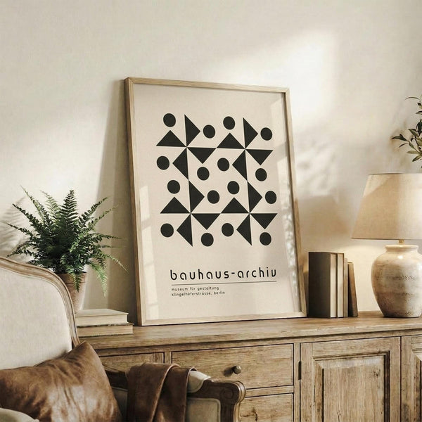 May include: Black and white geometric poster with circles and triangles in Bauhaus style, displayed in light wood frame on rustic wooden sideboard. Room setting shows cream-colored armchair with brown leather cushion, small potted fern, ceramic vase, and stacked books against white wall with natural lighting.