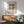 May include: Framed Bauhaus-style poster with black geometric pattern of triangles and circles on cream background, displayed above mid-century modern wood credenza with white ceramic lamp, next to sage green upholstered armchair with wooden frame in sunlit room with white walls and hardwood floors.