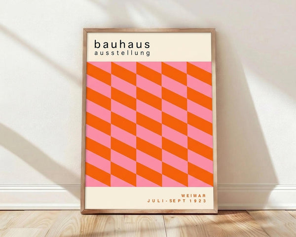 May include: Framed poster with geometric pink and orange cube pattern leaning against white wall on light wood floor. Clean minimalist room setting with natural shadows. Wooden frame surrounds cream-colored print featuring "bauhaus ausstellung" text above repeating 3D cubic design in bright coral and rose pink tones, with "Weimar Juli-Sept 1923" at bottom.