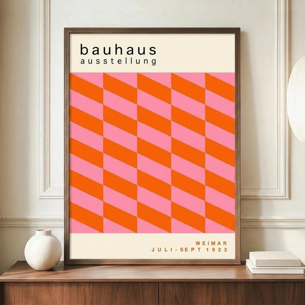 May include: Framed Bauhaus exhibition poster with geometric pink and orange cube pattern displayed on wooden sideboard next to white ceramic vase and stacked books in elegant room with white paneled walls
