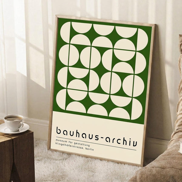 May include: Framed green and cream geometric poster with circular patterns leaning against white wall corner. Natural wood frame, cream mat border with black text below the design. Small wooden side table with white coffee cup and saucer nearby. Beige knit throw blanket visible in corner on white fluffy rug.