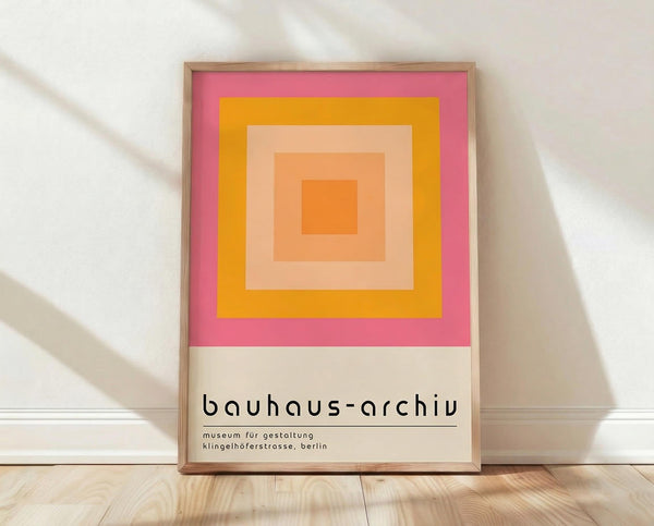 May include: Framed geometric poster with concentric squares in warm tones - pink outer border, golden yellow, peachy salmon, and bright orange center square. Text reads "bauhaus-archiv museum für gestaltung klingelhöferstrasse, berlin" in lowercase modern font. Natural wood frame sits on light wooden floor against white wall with soft shadow patterns.