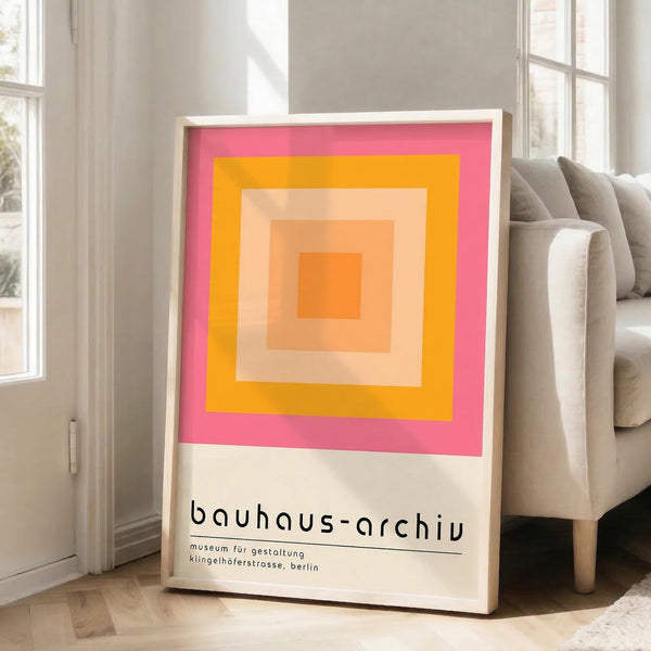 May include: Framed poster with concentric squares in gradient colors from pink outer border through orange and yellow to peach center, displayed against white wall next to beige upholstered sofa in bright living room with large windows and natural lighting on wooden floors.