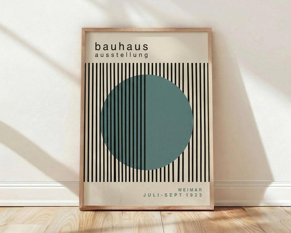 May include: Framed Bauhaus exhibition poster in light wood frame leaning against white wall on wooden floor. Design features black and white vertical stripes with large teal green circle overlay. Text reads "bauhaus ausstellung" at top and "WEIMAR JULI-SEPT 1923" at bottom on cream background. Minimalist geometric modernist style typical of 1920s Bauhaus movement. Natural lighting creates soft shadows on wall.