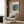 May include: Minimalist framed poster with black vertical stripes and teal circle mounted on wood wall panels above beige upholstered armchair. Modern reading corner features brass floor lamp with white marble base and dark side table with open book. Neutral tones throughout with geometric patterned throw pillow and sheer curtains creating warm natural lighting in contemporary interior setting.