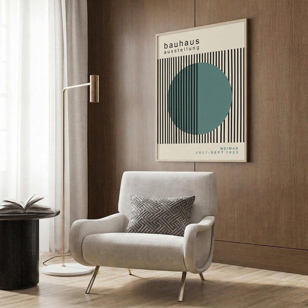 May include: Minimalist framed poster with black vertical stripes and teal circle mounted on wood wall panels above beige upholstered armchair. Modern reading corner features brass floor lamp with white marble base and dark side table with open book. Neutral tones throughout with geometric patterned throw pillow and sheer curtains creating warm natural lighting in contemporary interior setting.