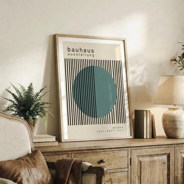 May include: Framed Bauhaus exhibition poster with black and white vertical stripes and teal circle, displayed on rustic wooden sideboard alongside fern plant, ceramic vase, and books in warm neutral living room setting with beige armchair