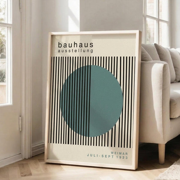 May include: Framed Bauhaus exhibition poster with black and white vertical stripes and teal circle, displayed on wooden floor beside beige sofa in bright living room with white walls and windows