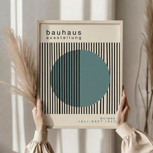May include: Person in beige sweater holding framed Bauhaus exhibition poster featuring teal green circle overlaying black and white vertical stripes on cream background, displayed against soft neutral curtains with dried pampas grass visible to the left