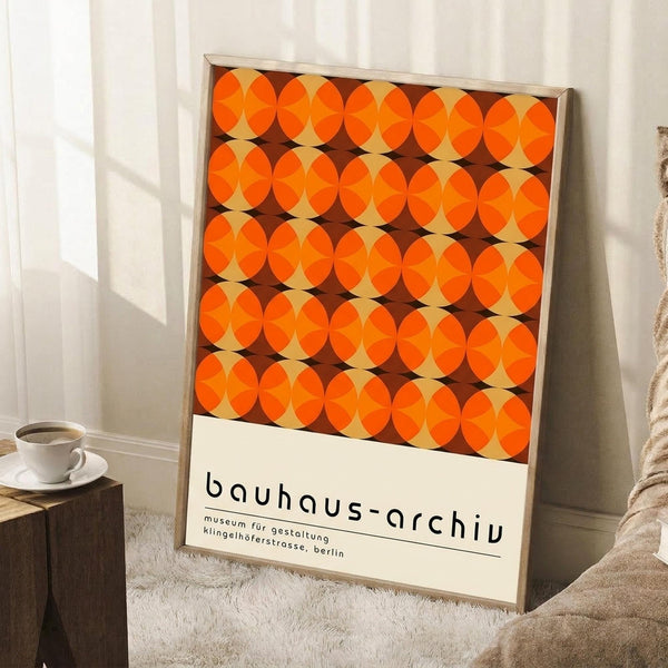May include: Framed poster with geometric orange and brown circular pattern leaning against white wall corner. Modern minimalist room setting with white coffee cup on wooden block side table and beige throw blanket. Natural lighting creates soft shadows on white walls and light wooden floors.