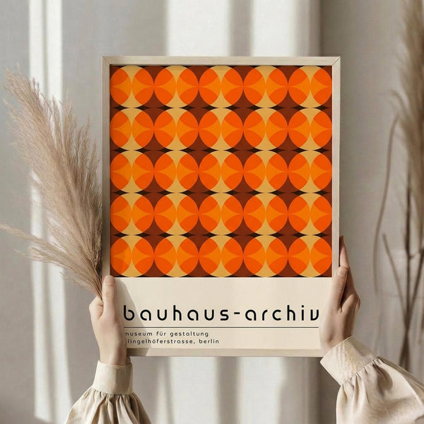 May include: Person holding framed retro geometric poster with repeating orange and brown circles pattern in warm sunlit room with dried pampas grass and white curtains. The minimalist wooden frame contains vintage Bauhaus-style artwork with overlapping circular motifs creating diamond shapes between them.